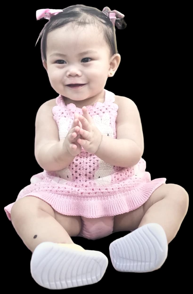 Lili, sitting and clapping in a pink knitted dress with bunny motifs and pink bows in her hair
