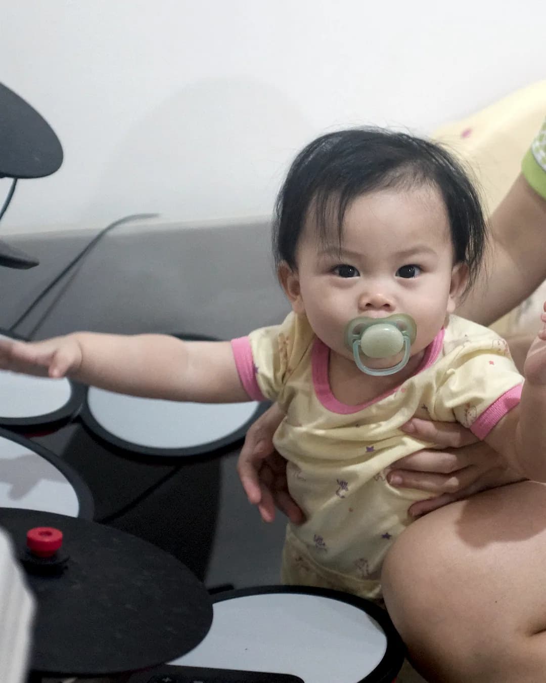 Lili sitting up at an electronic drum kit with a pacifier, one arm stretched out